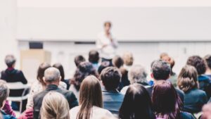 大学の様なtころで教授の様な人がホワイトボードの前で話している。大勢の人が聞いている